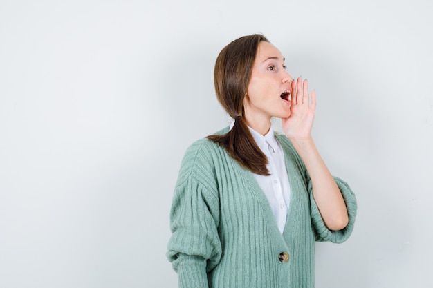 Young woman in blouse, cardigan shouting or announcing something and looking focused , front view.