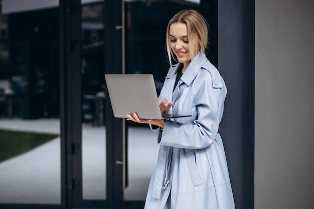 Young woman in blue coat standing by office building holding laptop