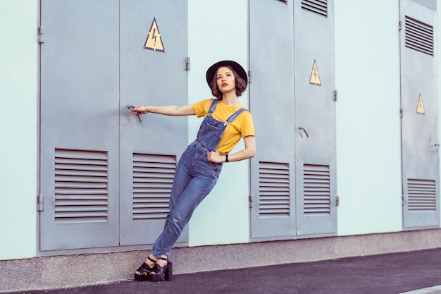Young woman in blue denim overalls and yellow tshirt with black hat sensual looking up while posing near industrial building . outdoor shot in the summertime