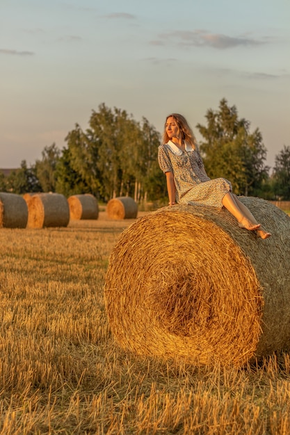 Foto giovane donna in abito blu da vicino sullo sfondo del campo con mucchi di fieno all'ora del tramonto.