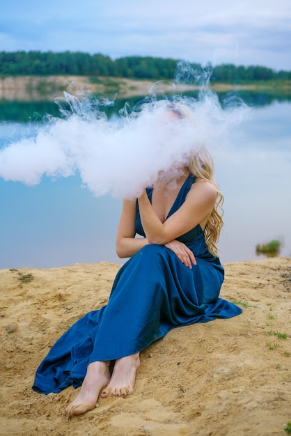A young woman in a blue dress sits on the shore of a lake during the day. A cloud of smoke before