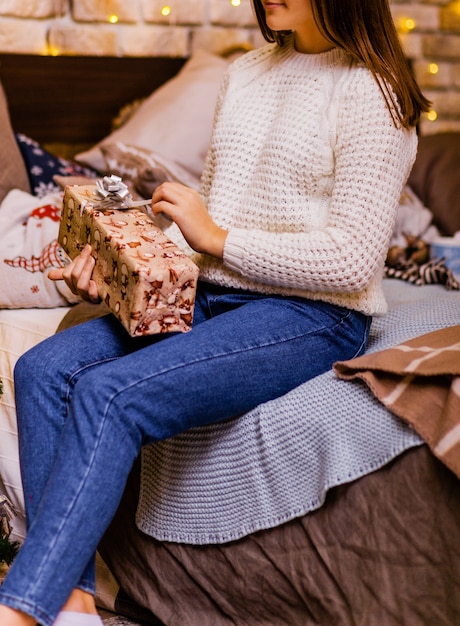 A young woman in blue jeans and a white knitted sweater sits on the bed and opens a New Year gift. Photo