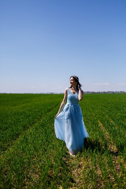 Young woman in a blue long dress on a background of green field. Fashion portrait of a beautiful girl with a smile on her face