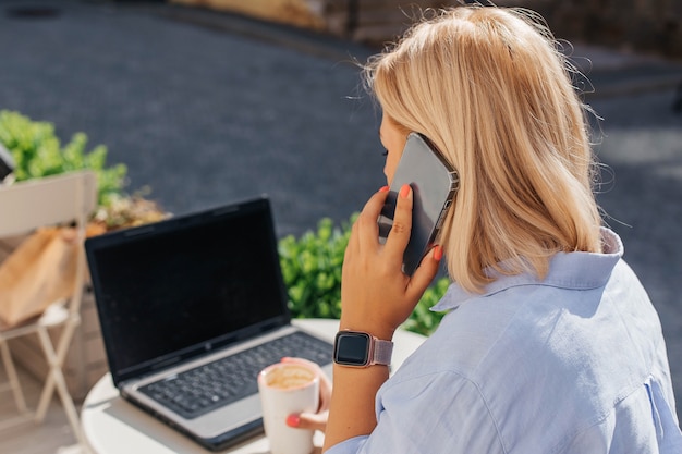 Young woman in a blue shirt is working on a laptop in a cafe and talking on the phone