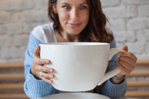 Young woman in blue sweater frowning with head in hand and holding giant cup of tea at home. Woman is feeling headache, holding big cup hot drink. Concept of disease, sickness and illness. Copy space