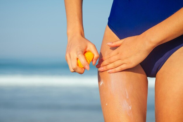 Young woman in a blue swimsuit holding a plastic orange package\
spray bottle and putting sunscreen on her body summer sun\
ultraviolet protection spf on the beach