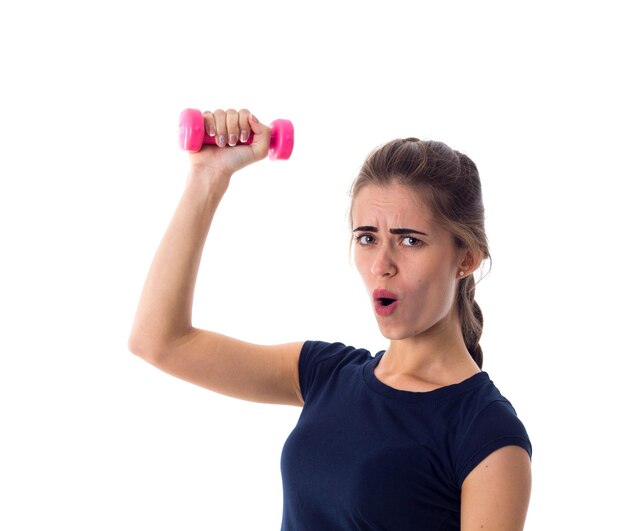 Молодая женщина в синей футболке с розовой гантелью в руке на белом фоне в студии