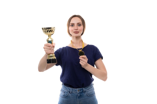 A young woman in a blue tshirt and jeans poses with the winner's cup and gold medal on a white background The concept of sports competitions