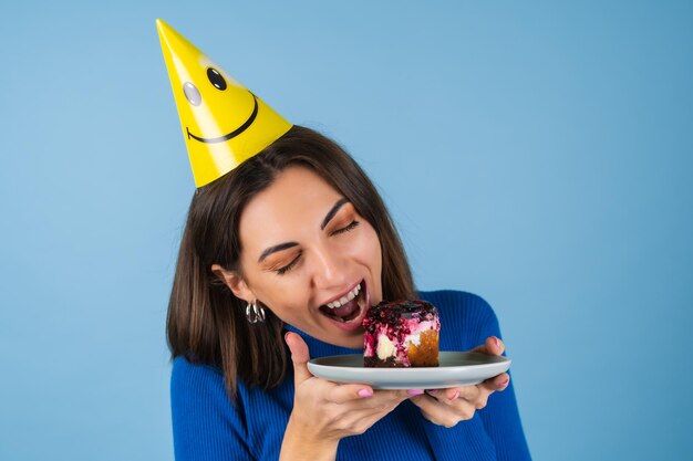 Young woman on a blue wall celebrates a birthday, holds a piece of cake, happy, bites the cake