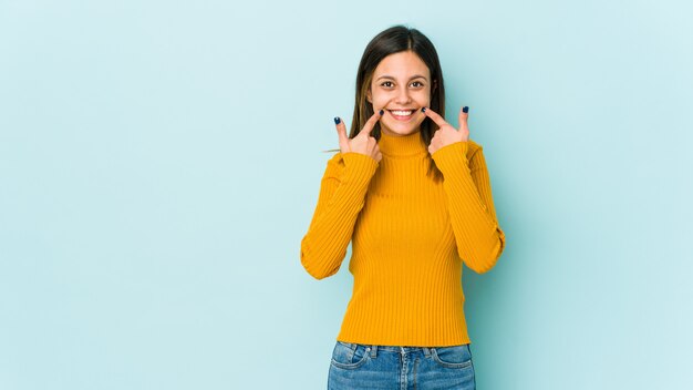 Young woman on blue wall smiles, pointing fingers at mouth.