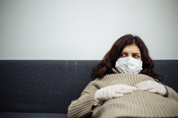 Photo young woman bored sitiing home during quarantine due to coronavirus pandemia. beautiful girl stays home wearing medical mask and gloves laying on a sofa. covid-19 epidemia worldwide concept.