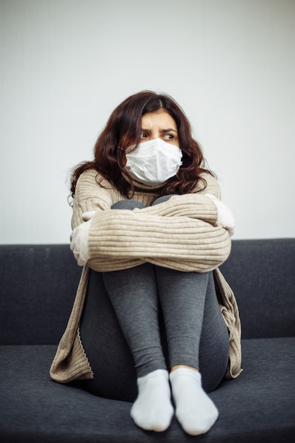 Young woman bored sitiing home during quarantine due to coronavirus pandemia. Beautiful girl stays home wearing medical mask and gloves laying on a sofa. Covid-19 epidemia worldwide concept.