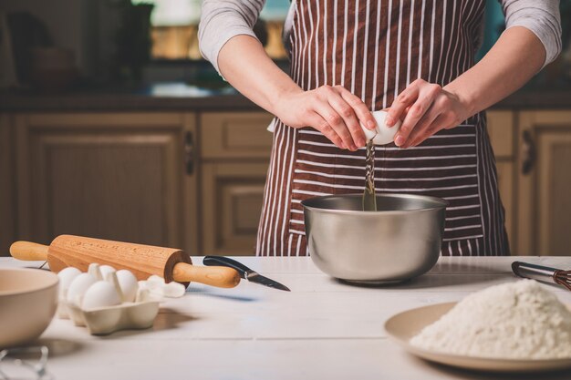 Young woman breaking egg over bowl with dough, close-up. A woman in a striped apron is cooking in the kitchen