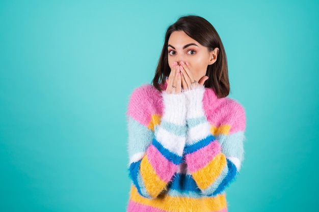 Photo young woman in a bright multicolored sweater on blue in shock covers her mouth with her palms