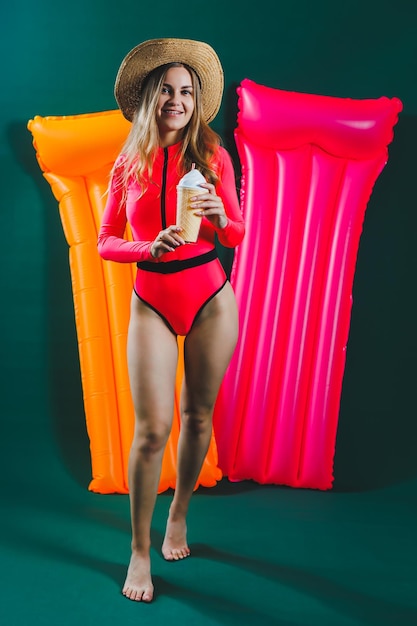 A young woman in a bright pink swimsuit and hat with an\
inflatable pink mattress on a green background summer rest girl\
with a cocktail in her hands in a bathing suit