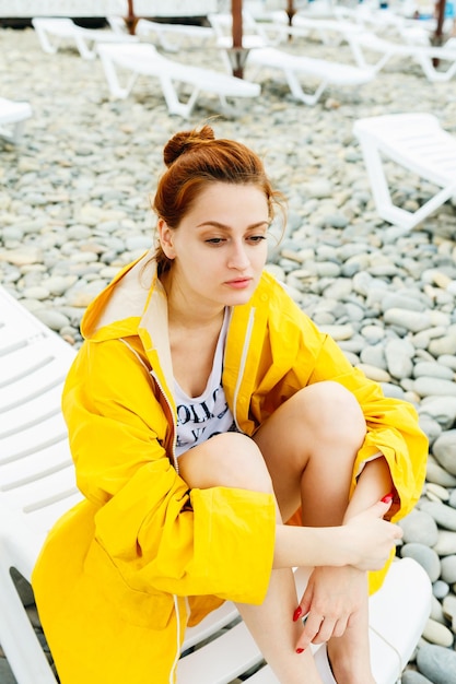 Young woman in bright raincoat sitting on sunbed on pebble beach