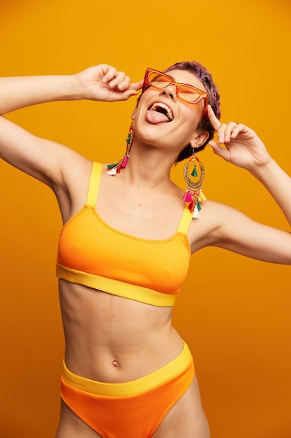 밝은 노란색 피트니스 운동복을 입은 젊은 여성은 배꼽이 열려 있고 주황색 배경에 선글라스가 웃고 있습니다.