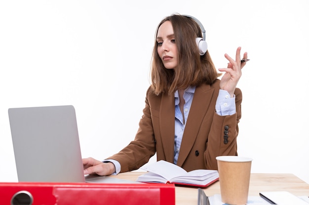 A young woman in a brown business suit is working at a laptop with headphones on her head at the workplace