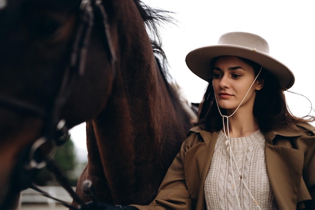 馬の近くに茶色のコートと帽子をかぶった若い女性の高画質写真