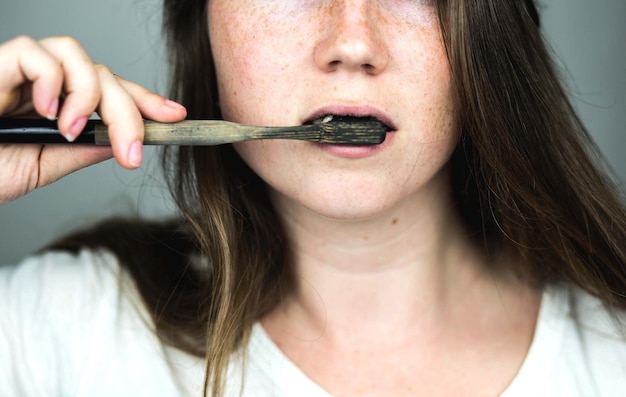 歯のホワイトニングのために白い背景に活性炭と黒い歯ブラシを使った黒い歯磨き粉で歯を磨く若い女性