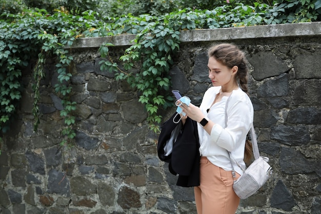 Young woman in business clothes stands outdoors near retaining wall and looks at the smartphone