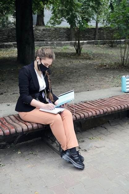 Young woman in business suit and facial mask sits on bench outdoors