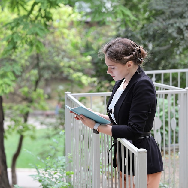 Young woman in business suit stands near the railing in the park and reading on tablet