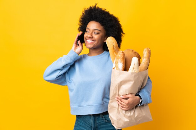 Young woman buying something bread isolated on yellow keeping a conversation with the mobile phone