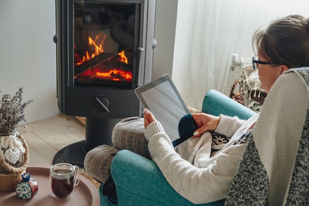 Photo young woman by the fireplace, sitting in a cozy armchair, with a warm blanket, using a tablet