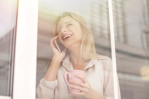 Young woman in cafe standing near window having phone call drinking tea joyful