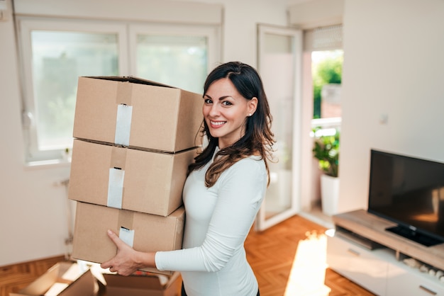 Young woman carrying cardboard boxes indoors. Smiling and looking at camera.