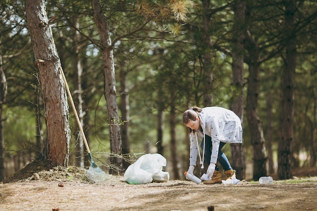 Young woman in casual clothes, gloves cleaning rubbish into trash bags in park or forest on green wall