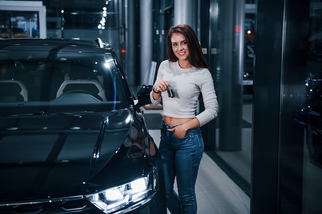 Young woman in casual clothes standing near modern black car with keys in hands.