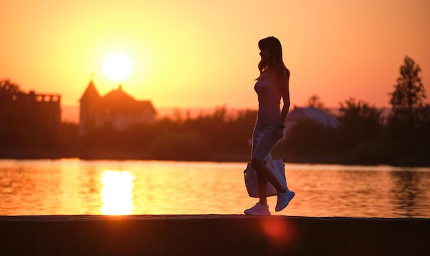 따뜻한 저녁 여름 휴가와 여행 컨셉에 호숫가를 걷고 있는 캐주얼 복장을 한 젊은 여성