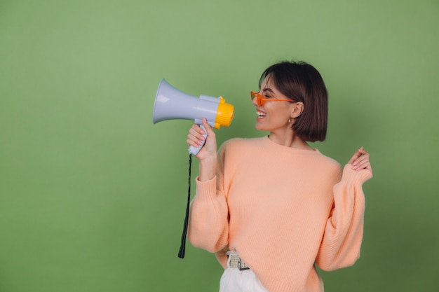 Young woman in casual peach and orange eyeglasses sweater isolated on green olive wall happy screaming in megaphone copy space