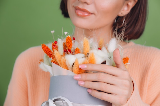 Photo young woman in casual peach sweater  isolated on green olive wall  hold  orange white flower box composition of cotton flowers gypsophila wheat and lagurus for a gift happy amazed surprised