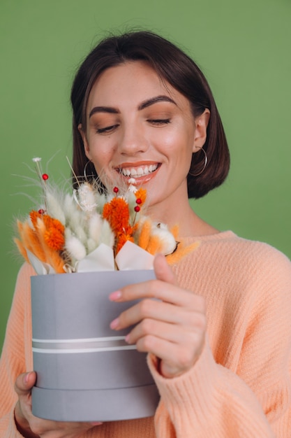 緑のオリーブの壁に分離されたカジュアルな桃のセーターの若い女性は、ギフトのための綿の花カスミソウ小麦とラグラスのオレンジ色の白いフラワーボックスの構成を保持します幸せな驚き驚き