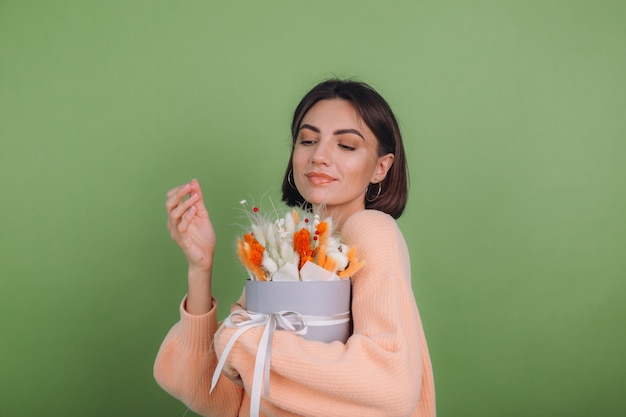 緑のオリーブの壁に分離されたカジュアルな桃のセーターの若い女性は、ギフトのための綿の花カスミソウ小麦とラグラスのオレンジ色の白いフラワーボックスの構成を保持します幸せな驚き驚き