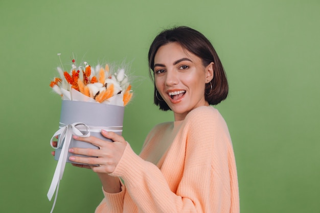 Young woman in casual peach sweater  isolated on green olive wall  hold  orange white flower box composition of cotton flowers gypsophila wheat and lagurus for a gift happy amazed surprised