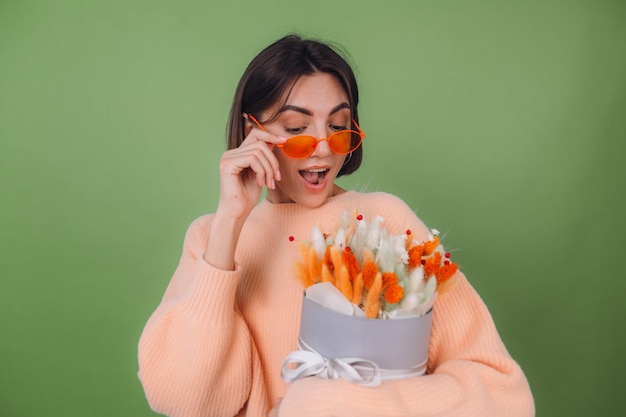 Young woman in casual peach sweater  isolated on green olive wall  hold  orange white flower box composition of cotton flowers, gypsophila, wheat and lagurus for a gift happy surprised