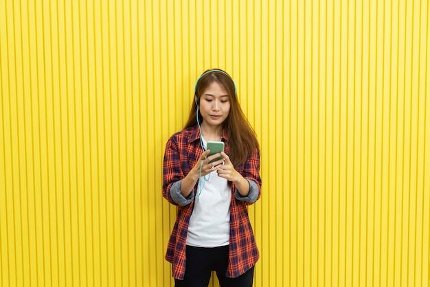 Young woman in casual using mobile and listening music on yellow wall.