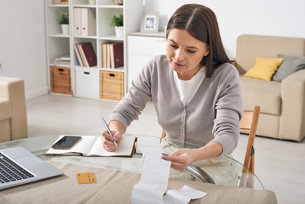 Young woman in casualwear looking through payment receipts and writing down the numbers in notebook