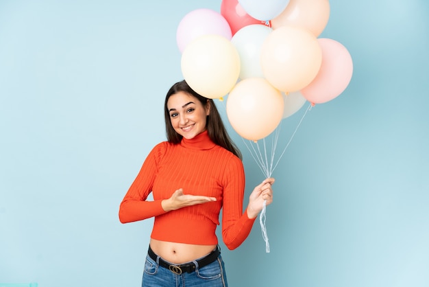 Young woman catching many balloons on blue extending hands to the side for inviting to come