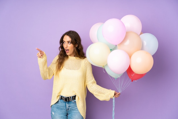 Young woman catching many balloons over isolated on purple wall pointing away