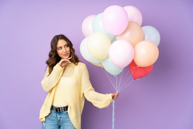 Young woman catching many balloons over isolated on purple wall thinking an idea