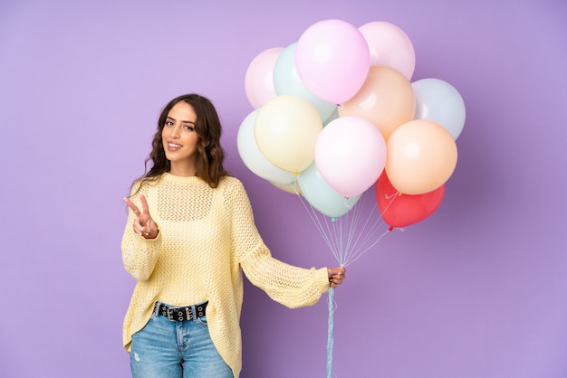 Young woman catching many balloons over on purple wall smiling and showing victory sign