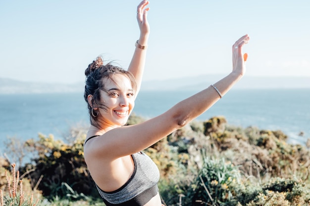 Young woman celebrates finishing a outdoor exercise session on\
the coastline dancing and raising arms to the air happy young\
people training during a sunny summer day preparing for the beach\
body
