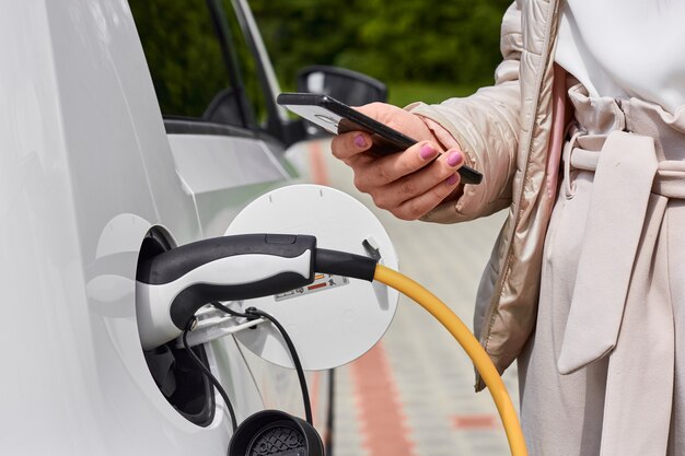 Giovane donna che carica un'auto elettrica alla stazione di ricarica pubblica e paga utilizzando un telefono cellulare. veicolo ecologico innovativo.