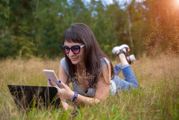 若い女性はスマートフォンをチェックし、屋外の夏の芝生でラップトップで作業します