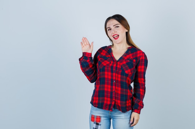 Young woman in checked shirt, jeans greeting with open hand and looking confident , front view.